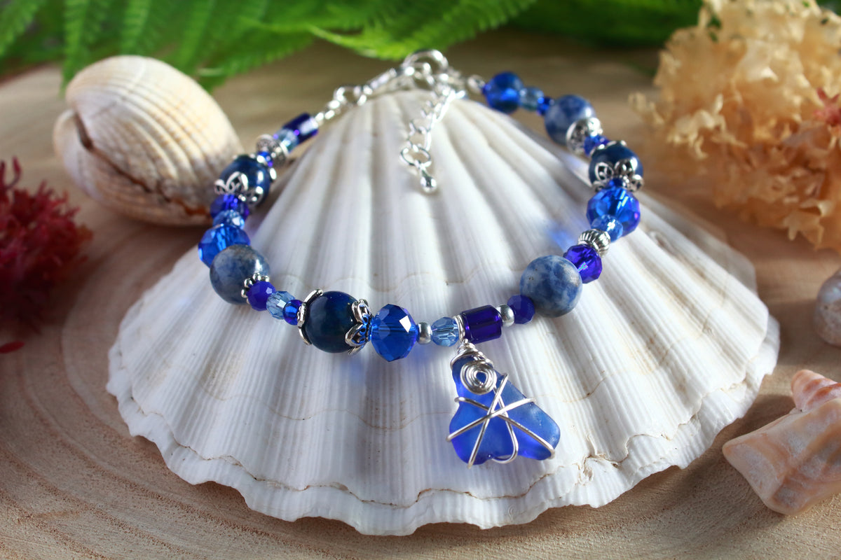 A boho beaded sea glass bracelet with blue and silver beads plus blue sea glass wrapped in silver wire, displayed on a white seashell with decorative shells and greenery in the background.