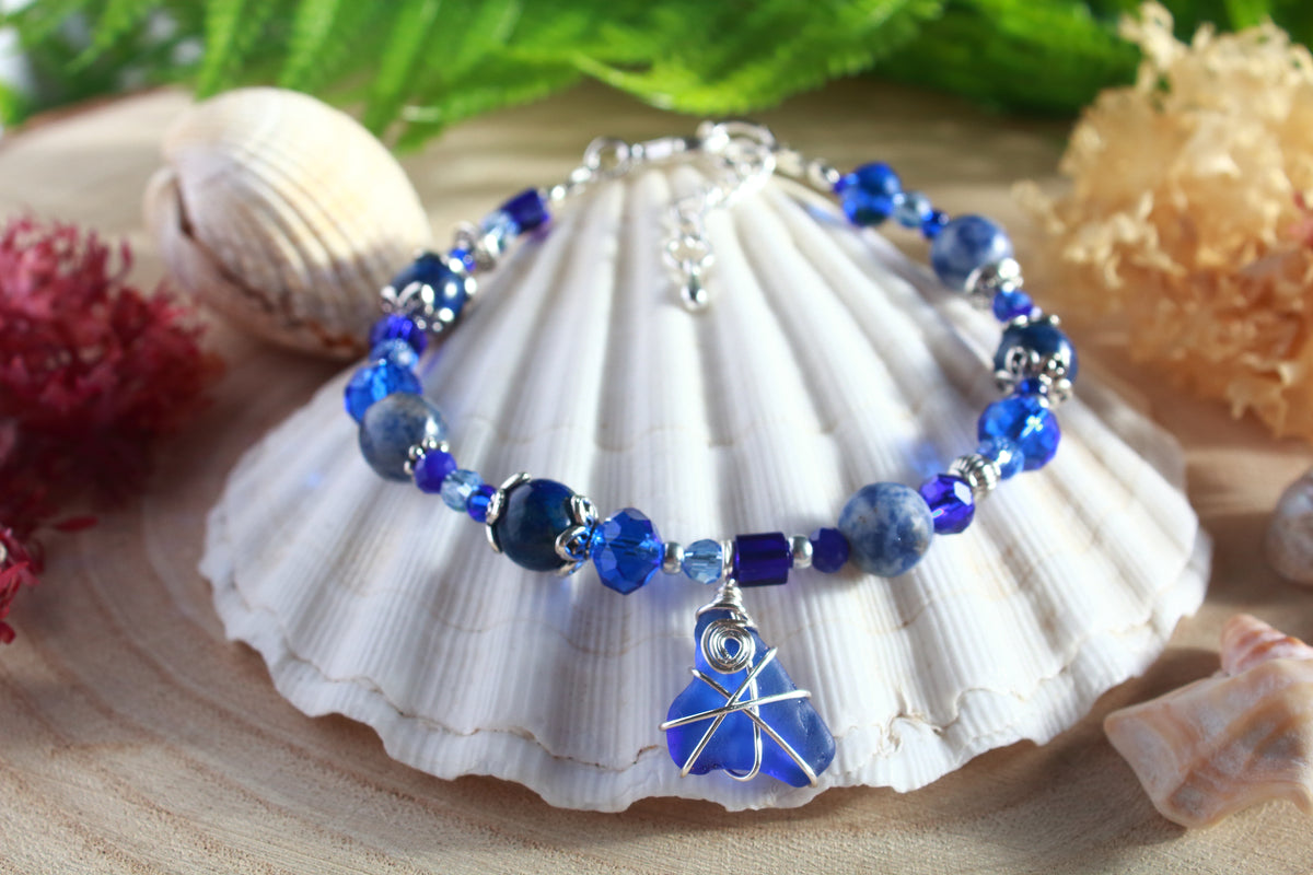 A boho beaded sea glass bracelet with blue and silver beads plus blue sea glass wrapped in silver wire, displayed on a white seashell with decorative shells and greenery in the background.